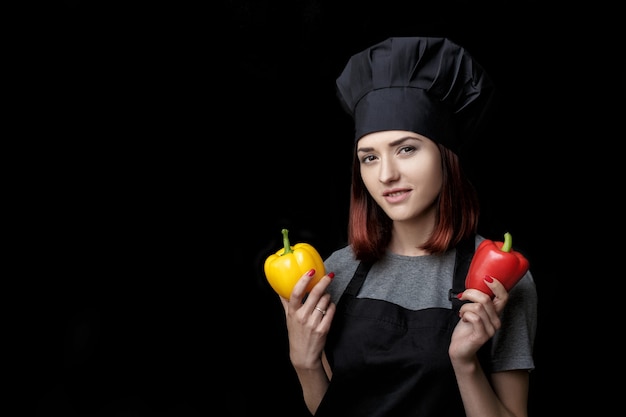 Jonge aantrekkelijke vrouw chef-kok in zwart uniform houdt paprika op zwarte achtergrond