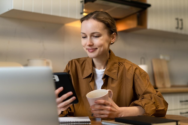 Jonge aantrekkelijke vrouw die lacht en gadgets gebruikt, telefoon en laptop voor communicatie op afstand terwijl ze in de moderne keuken zit