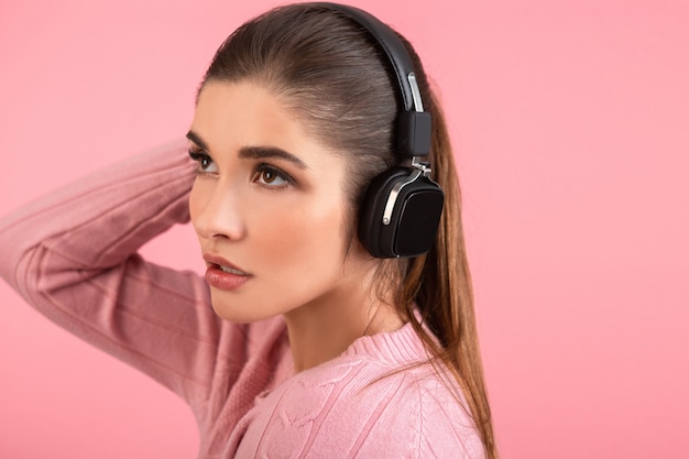 Jonge aantrekkelijke vrouw die naar muziek luistert in een draadloze koptelefoon met een roze trui die lacht en een vrolijke positieve stemming op een roze achtergrond