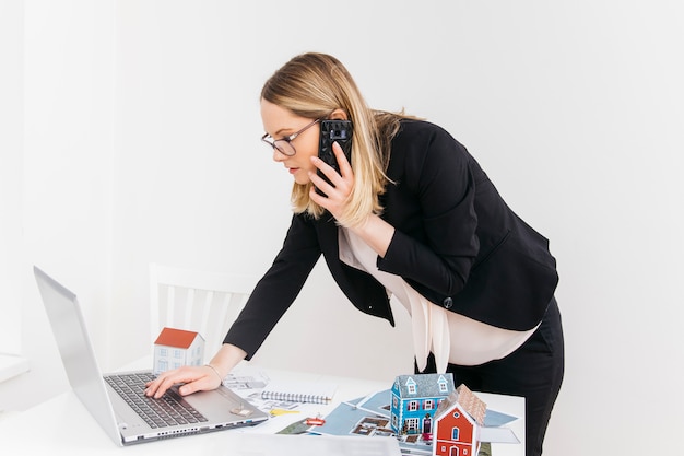 Foto jonge aantrekkelijke vrouw die op cellphone spreekt terwijl het werken aan laptop in onroerende goederenbureau