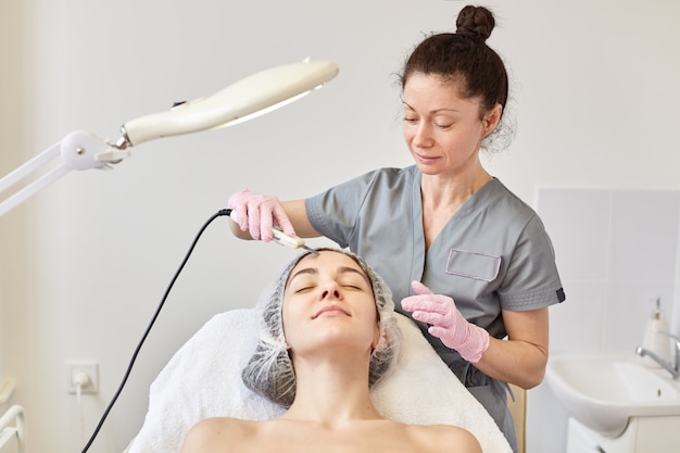 Jonge aantrekkelijke vrouw die ultrasone gezichtshuid reinigende behandeling krijgen door professionele schoonheidsspecialist