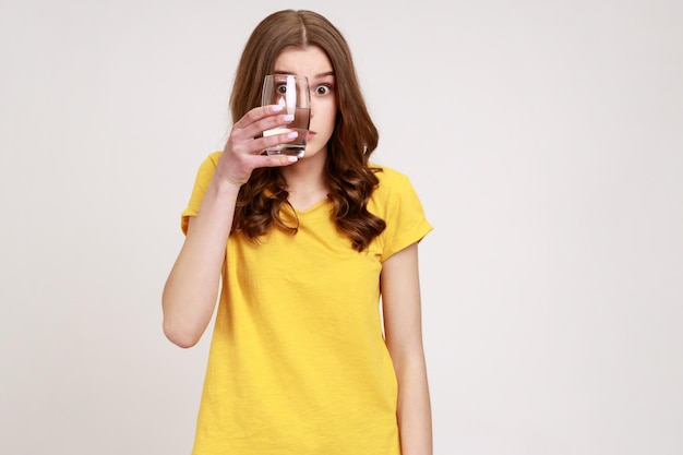 Jonge aantrekkelijke vrouw draagt gele casual T-shirt die oog bedekt met glas water op zoek naar glas met grote verbaasde ogen Indoor studio-opname geïsoleerd op een grijze achtergrond