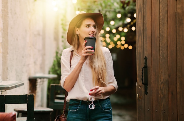 Jonge aantrekkelijke vrouw in casual stijl kleding wandelen langs de binnenplaats van de oude stad met een kopje koffie onderweg