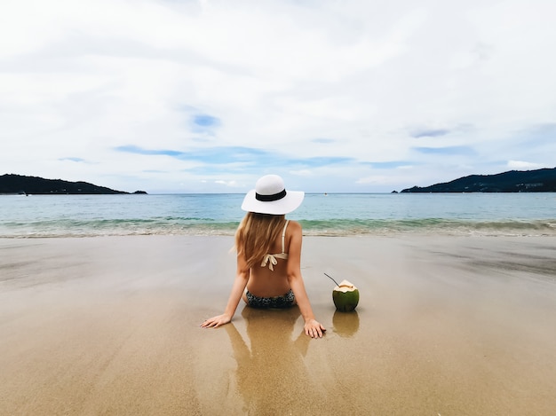 Jonge aantrekkelijke vrouw in een hoed op de kust van de blauwe overzeese leunend met een kokosnoot, Thailand