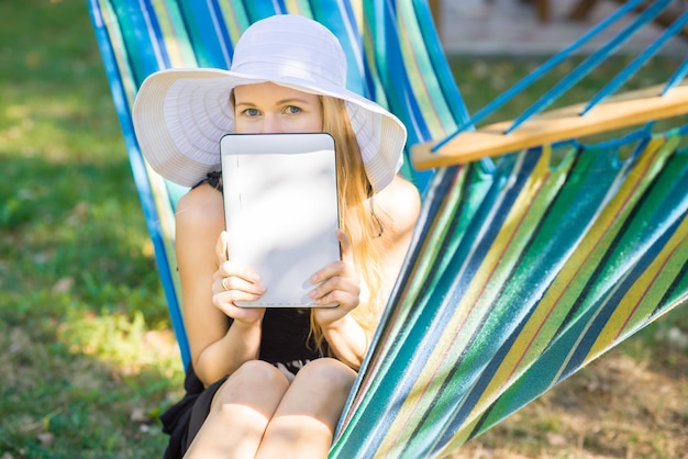 Jonge aantrekkelijke vrouw in hoed met behulp van een tablet op de hangmat in de tuin