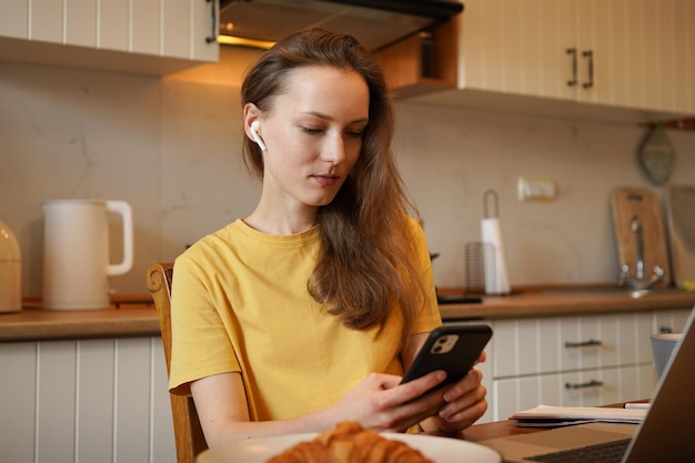 Jonge aantrekkelijke vrouw in koptelefoon en geel t-shirt tekst aan het typen op de telefoon terwijl ze thuis aan tafel zit