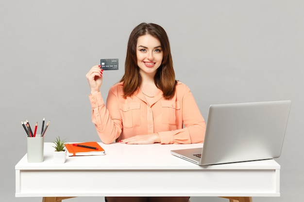 Jonge aantrekkelijke vrouw in pastel casual kleding met creditcard zitten werk aan bureau met pc laptop geïsoleerd op een grijze achtergrond. Prestatie zakelijke carrière levensstijl concept. Bespotten kopie ruimte.