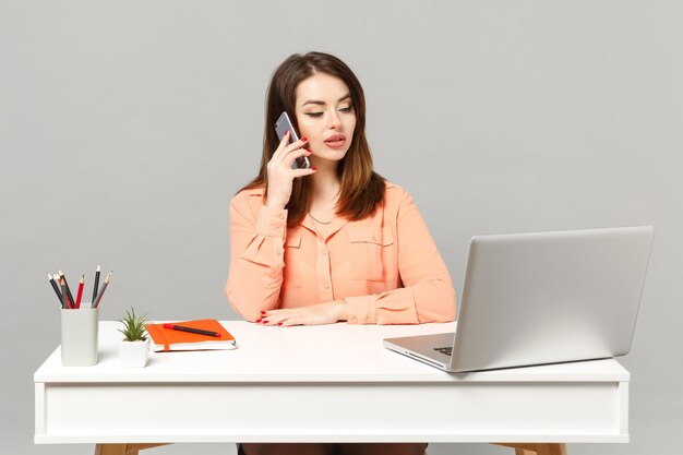 Jonge aantrekkelijke vrouw in pastel casual kleding praten op mobiele telefoon, zitten werk aan bureau met pc-laptop geïsoleerd op een grijze achtergrond. Prestatie zakelijke carrière levensstijl concept. Bespotten kopie ruimte.