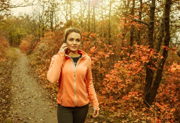 Jonge aantrekkelijke vrouw in sportkleding die aan de telefoon praat terwijl ze langs een pad loopt met rode herfstbladeren in het bos