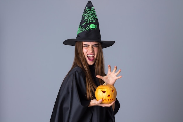 Jonge aantrekkelijke vrouw in zwart kostuum van heks met zwarte hoed die een pompoen in studio houdt