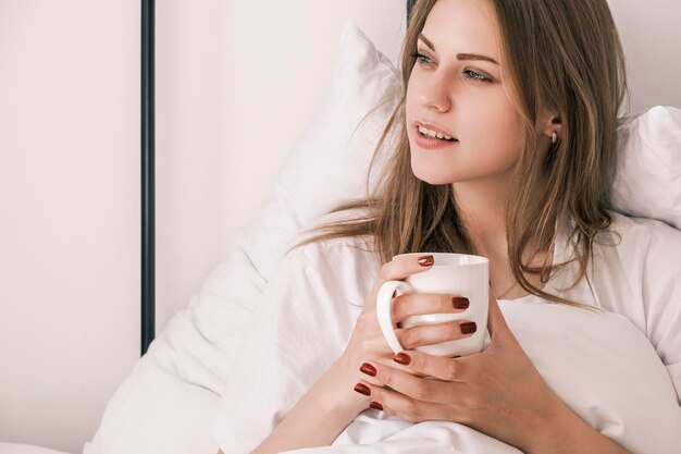 Jonge aantrekkelijke vrouw ligt in bed na het wakker worden met een witte kop in haar hand en kijkt uit het raam