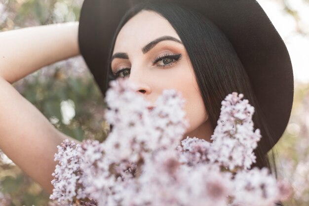 Jonge aantrekkelijke vrouw met mooie ogen in luxe elegante hoed bedekt gezicht met verse lila boeket bloemen in bloeiend park. Lief meisje geniet van de geur van lentebloesems en warme lentestralen zon