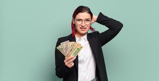 Jonge aantrekkelijke vrouw met rood haar die zich gestrest, bezorgd, angstig of bang voelt, met de handen op het hoofd, in paniek raakt bij vergissing. geld concept