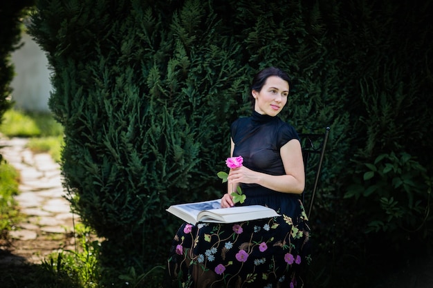 Jonge aantrekkelijke vrouw zit in een tuin met een groot boek en een roze bloem in haar handen