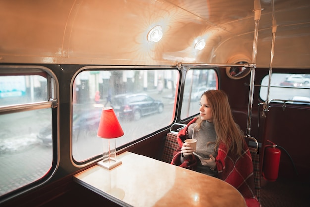 Jonge aantrekkelijke vrouw zittend aan de tafel in het buscafé is bedekt met een deken, houdt een kopje koffie in haar handen en kijkt in het raam