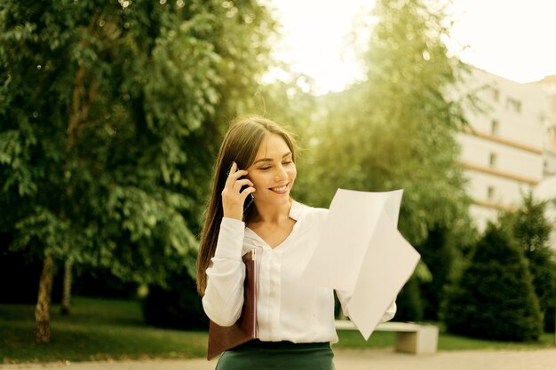 Jonge aantrekkelijke vrouwelijke manager houdt vellen papier buiten in de stad aan de telefoon. Alles in het bedrijfsleven