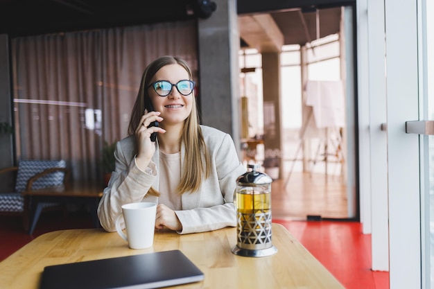 Jonge aantrekkelijke vrouwelijke manager praat op een mobiele telefoon en glimlacht terwijl ze alleen zit in de buurt van een groot raam in een café in haar vrije tijd en op een computer werkt Gelukkige vrouw rust in een café