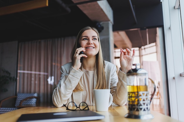 Jonge aantrekkelijke vrouwelijke manager praat op een mobiele telefoon en glimlacht terwijl ze alleen zit in de buurt van een groot raam in een café in haar vrije tijd en op een computer werkt Gelukkige vrouw rust in een café