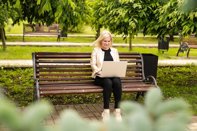 Jonge aantrekkelijke zakenvrouw die een pc-laptop gebruikt terwijl ze op een houten bankje in een stadspark zit.