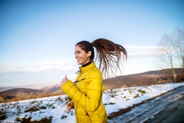 Jonge actieve atletische meisjesjogging in de wintersportkleding op sneeuw de winterweg met oortelefoons in de zonnige ochtend.