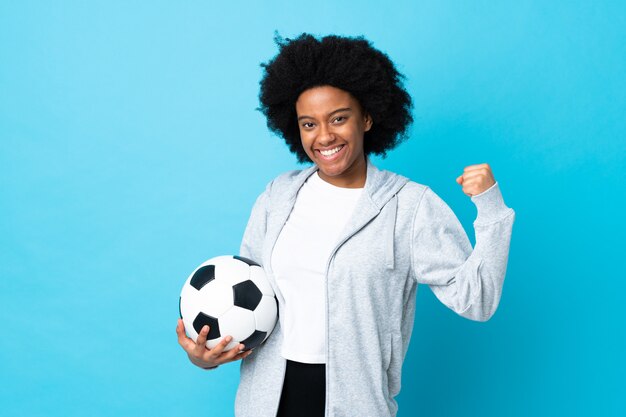 Jonge African American vrouw geïsoleerd op blauwe muur met voetbal vieren van een overwinning