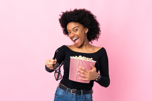 Jonge african american vrouw geïsoleerd op roze met 3d-bril en met een grote emmer popcorns