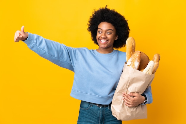 Jonge African American vrouw iets kopen brood geïsoleerd op geel met een duim omhoog gebaar