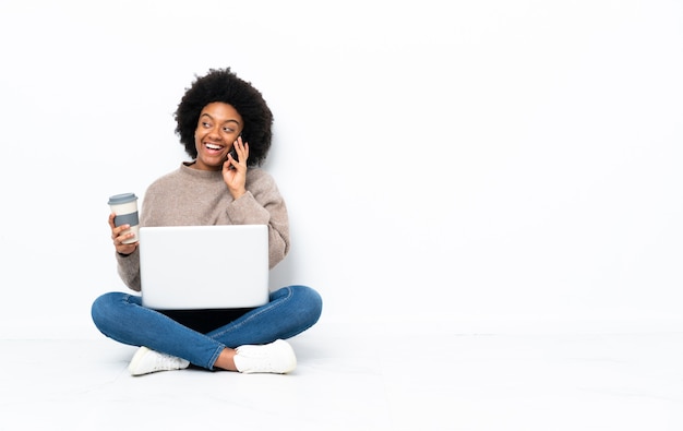 Jonge African American vrouw met een laptop zittend op de vloer met koffie om mee te nemen en een mobiel