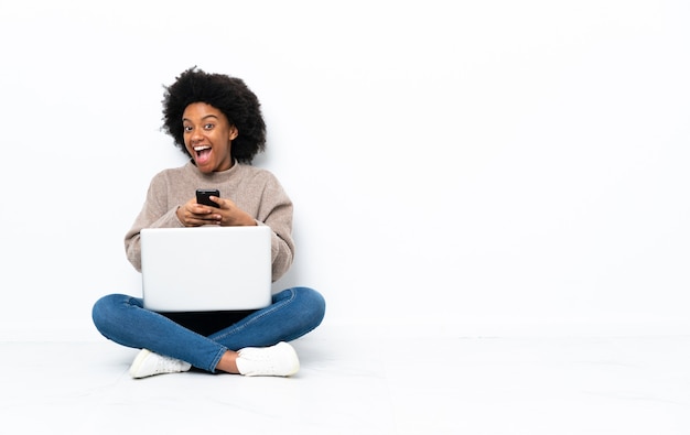 Jonge African American vrouw met een laptop zittend op de vloer verrast en een bericht verzenden
