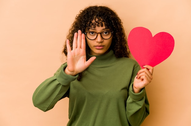 Jonge afrikaanse amerikaanse afrovrouw die een hart van de valentijnskaartendag houdt dat zich met uitgestrekte hand bevindt die stopbord toont, dat u verhindert.