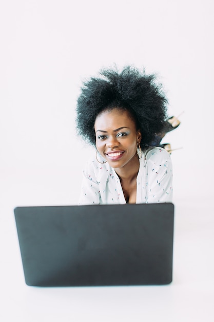 Jonge afrikaanse amerikaanse bedrijfsvrouw die laptop met behulp van, terwijl het liggen op de geïsoleerde vloer