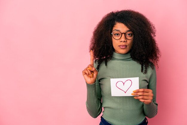 Jonge afrikaanse amerikaanse krullende vrouw die een hartsymbool op aanplakbiljet houdt dat nummer één met vinger toont.