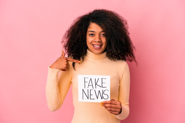 Jonge afrikaanse amerikaanse krullende vrouw die een vals nieuws op een aanplakbiljet houdt die met de hand naar de ruimte van een overhemds exemplaar wijst, trots en zelfverzekerd
