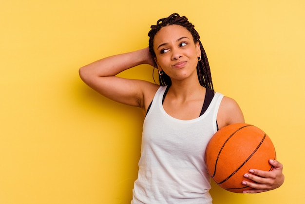 Jonge afrikaanse amerikaanse vrouw die basketbal speelt dat op gele muur wordt geïsoleerd die achterkant van het hoofd raakt, denkt en een keuze maakt.