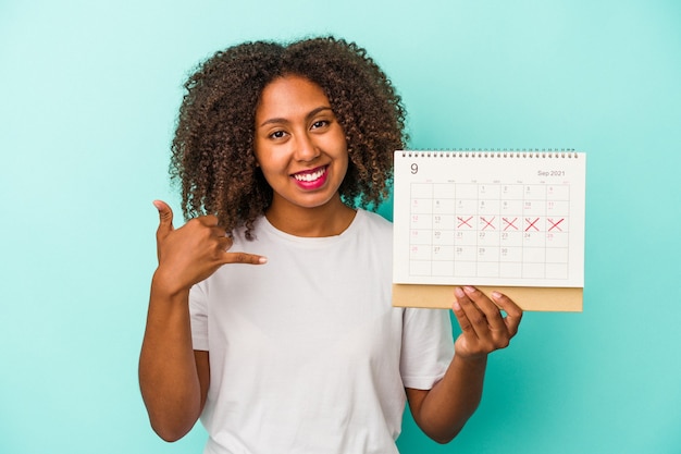 Jonge afrikaanse amerikaanse vrouw die een kalender houdt die op blauwe achtergrond wordt geïsoleerd die een mobiel telefoongesprekgebaar met vingers toont.