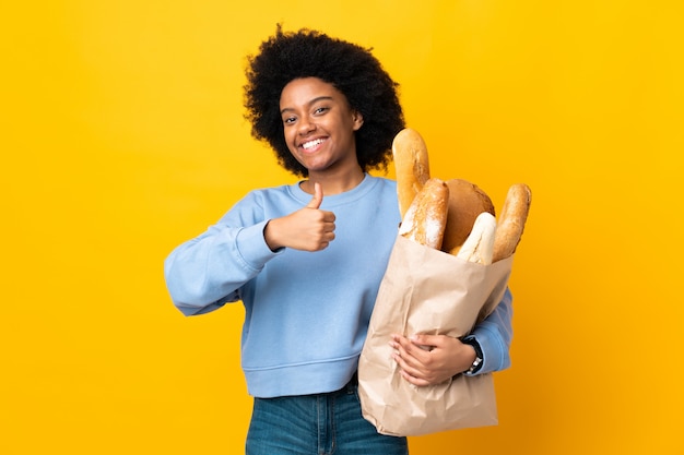 Jonge Afrikaanse Amerikaanse vrouw die iets brood koopt dat op gele achtergrond wordt geïsoleerd die duimen op gebaar geven