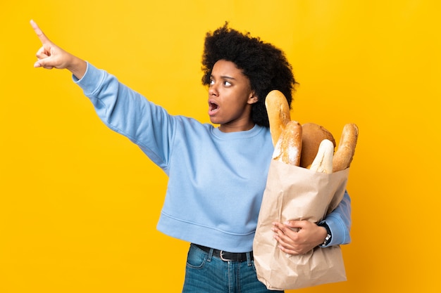 Jonge Afrikaanse Amerikaanse vrouw die iets brood koopt dat op gele achtergrond wordt geïsoleerd die weg richt