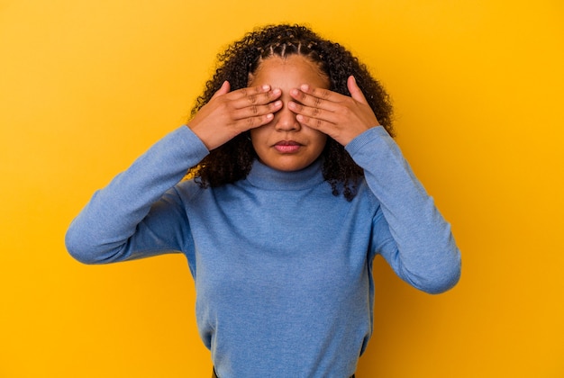 Jonge afrikaanse amerikaanse vrouw die op gele muur wordt geïsoleerd die bang is voor ogen met handen.