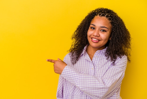 Jonge afrikaanse amerikaanse vrouw die op gele muur wordt geïsoleerd die opzij glimlacht richt en iets op lege ruimte toont.