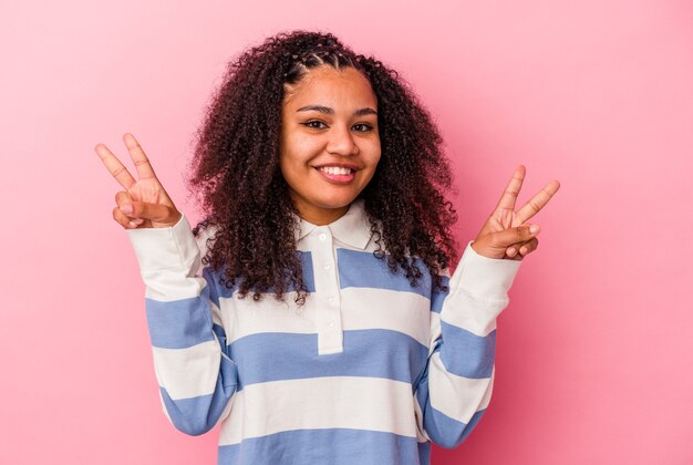 Jonge afrikaanse amerikaanse vrouw die op roze achtergrond wordt geïsoleerd die overwinningsteken toont en breed glimlacht.