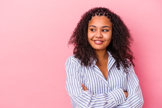 Jonge afrikaanse amerikaanse vrouw die op roze muur wordt geïsoleerd die zelfverzekerd met gekruiste wapens glimlacht.