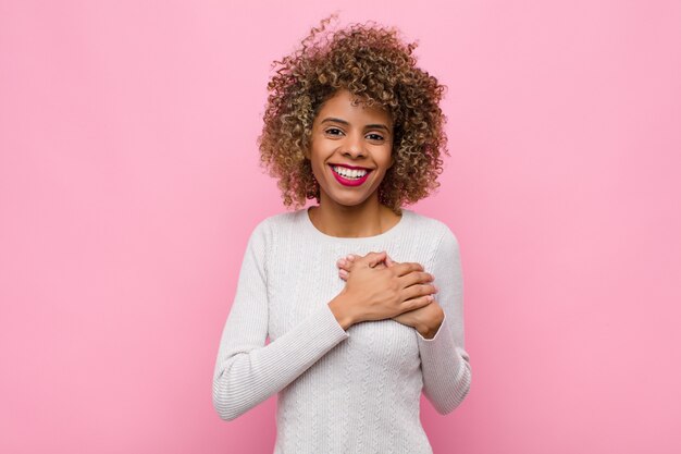 Jonge afrikaanse amerikaanse vrouw die zich romantisch, gelukkig en verliefd voelt, vrolijk lacht en hand in hand op roze muur dicht bij het hart houdt