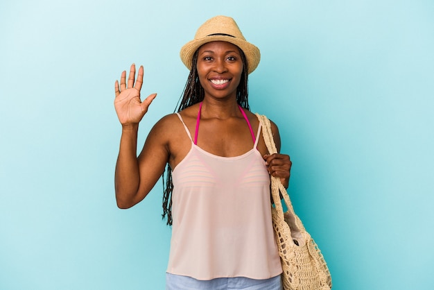 Jonge afrikaanse amerikaanse vrouw die zomerkleren draagt die op blauwe achtergrond worden geïsoleerd die vrolijk glimlachen die nummer vijf met vingers tonen.