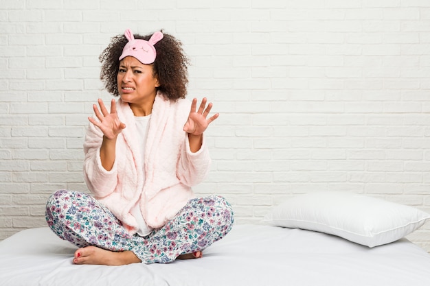 Jonge Afrikaanse Amerikaanse vrouw in het bed dat pijama draagt die iemand verwerpt die een gebaar van afschuw toont.