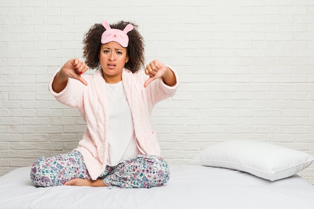 Jonge afrikaanse amerikaanse vrouw in het bed die pijama dragen die duim tonen en afkeer uitdrukken.