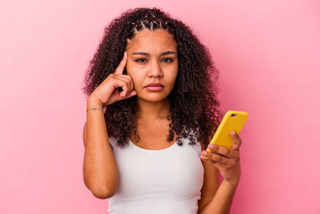 Jonge afrikaanse amerikaanse vrouw met een mobiele telefoon isoleerde een roze muur wijzende tempel met vinger, denkend, gericht op een taak.