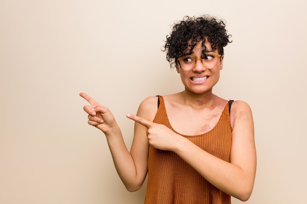 Jonge Afrikaanse Amerikaanse vrouw met huidgeboorteteken geschokt wijzend met wijsvingers aan een exemplaarruimte.