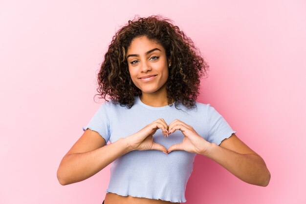 Jonge Afrikaanse Amerikaanse vrouw tegen een roze muur die en een hartvorm met handen glimlacht toont.
