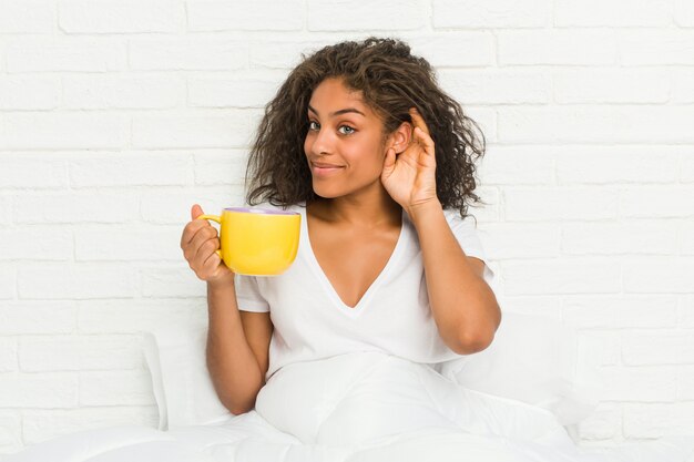 Jonge Afrikaanse Amerikaanse vrouwenzitting op het bed die een koffiemok houden die aan het luisteren van een roddel proberen.