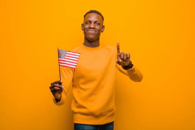 Jonge Afrikaanse Amerikaanse zwarte mens die tegen oranje muur een vlag van de VS houdt